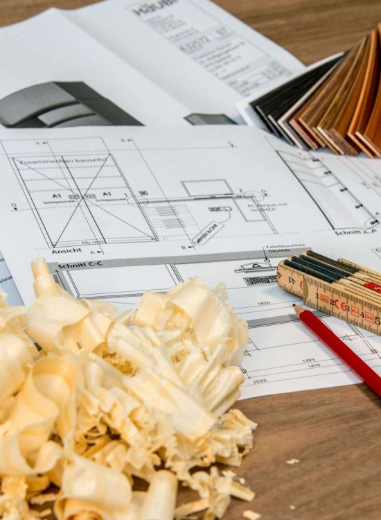 Holzspäne, technische Zeichnungen, Maßband, Bleistift und Farbproben auf einem Arbeitstisch in einer Schreinerei.