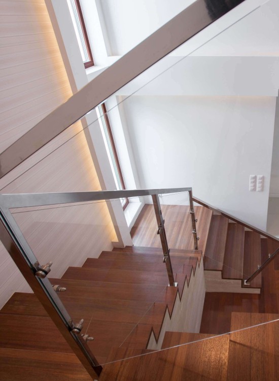 Moderne Holztreppe mit Glasgeländer und Edelstahlhandlauf in einem hellen Treppenhaus mit indirekter Beleuchtung.