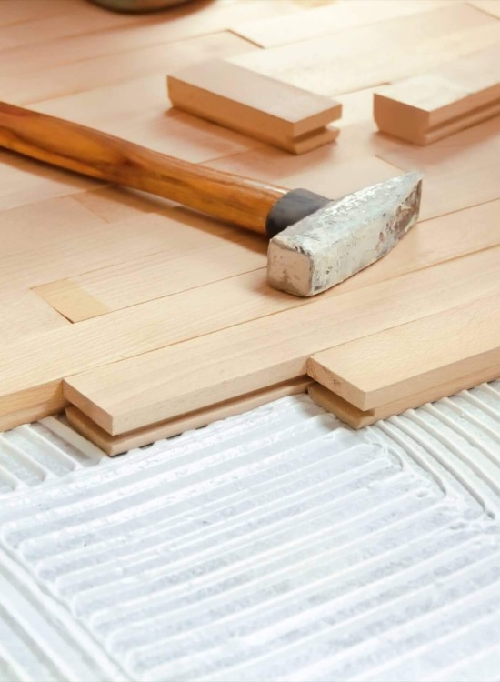 Verlegung eines hellen Parkettbodens, bei der Holzlamellen auf frischen Kleber gesetzt werden, ein Hammer liegt daneben.
