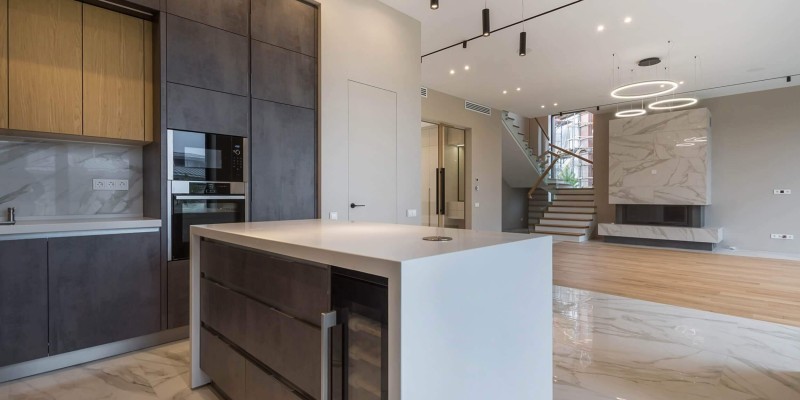 Offene, moderne Wohnküche mit Kochinsel, dunklen Fronten, Marmorfußboden und integriertem Kaminbereich mit Treppe im Hintergrund.